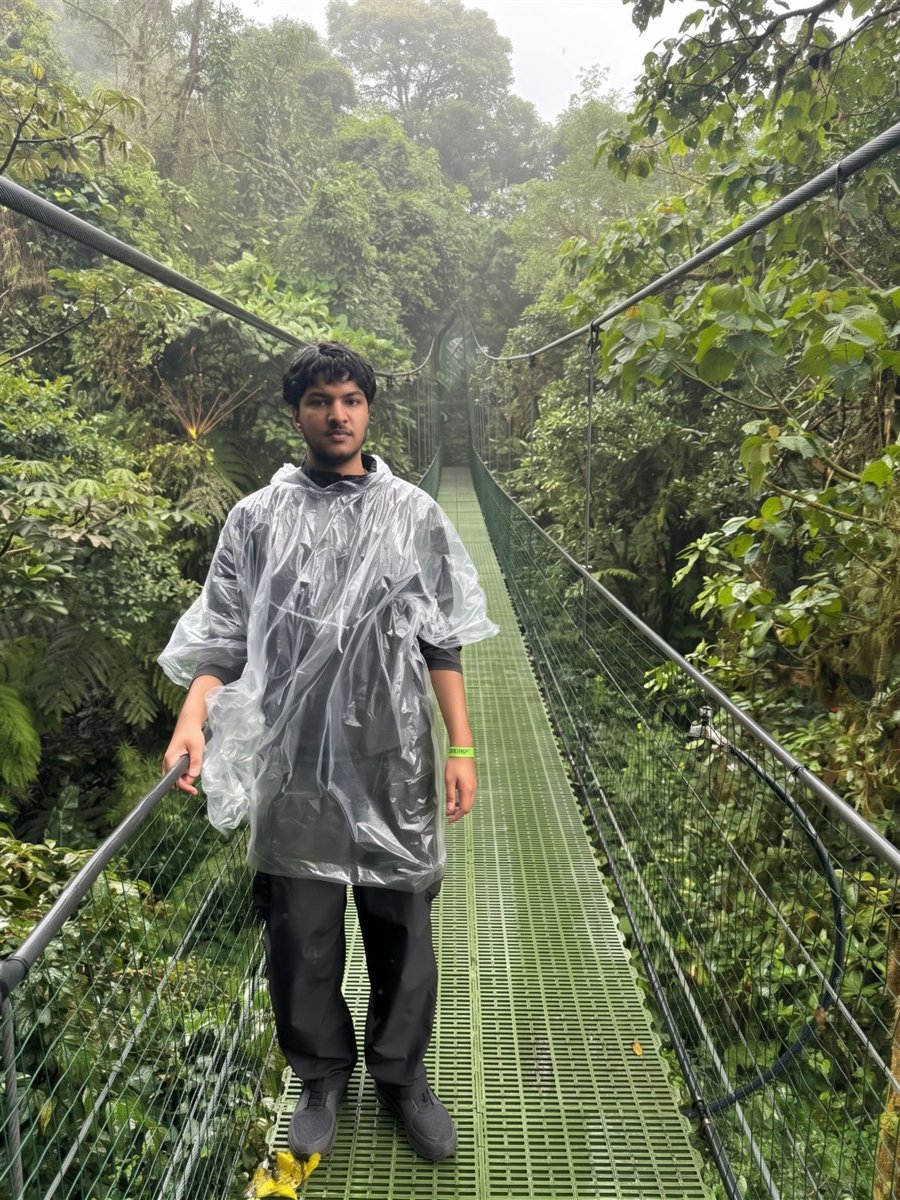 Ibrahim Raja in a clear rain poncho on a green suspension bridge surrounded by misty rainforest in Costa Rica